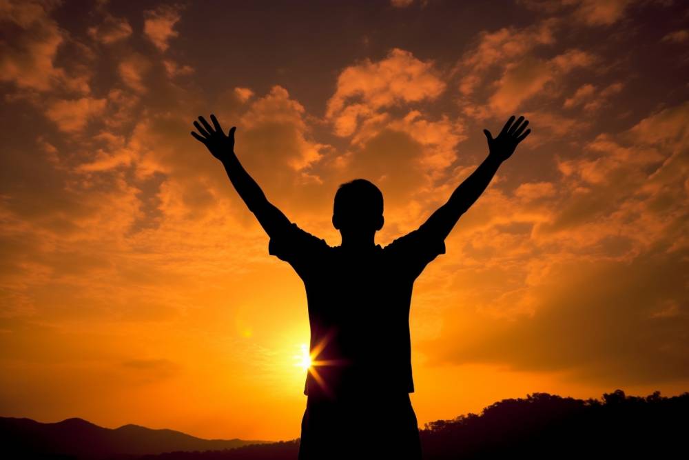Silhouette of man in the evening watching beautiful sunset with arms raised up outstretched cheering and happy